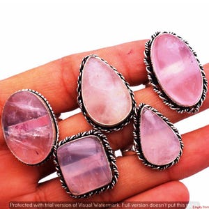 Könnte beinhalten: Fünf Ringe mit Rosenquarz-Edelsteinen in verschiedenen Formen: oval, tropfenförmig und quadratisch. Jeder Ring hat einen polierten rosa Stein, der in einer silberfarbenen, seiltexturierten Lünette gefasst ist. Die Ringe werden auf einer Hand präsentiert.