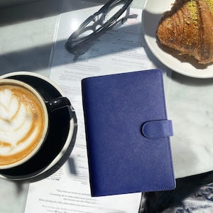 May include: A navy blue leather-bound planner with a snap closure sits on a marble table. A cup of coffee with latte art and a croissant are also on the table. The planner is closed and has a textured surface.