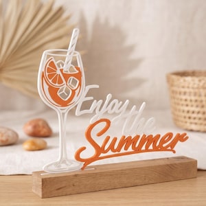 Könnte beinhalten: Dekoratives Holzschild mit den Worten "Enjoy the Summer" in Weiß und Orange. Das Schild zeigt ein Cocktailglas-Design mit Orangenscheiben und Eiswürfeln. Das Schild steht auf einem Holzsockel.