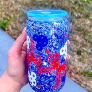 May include: A blue glitter tumbler with a matching blue lid. The tumbler is decorated with red bows, white clouds, blue Mickey Mouse silhouettes, and white stars. The tumbler is held by a person.