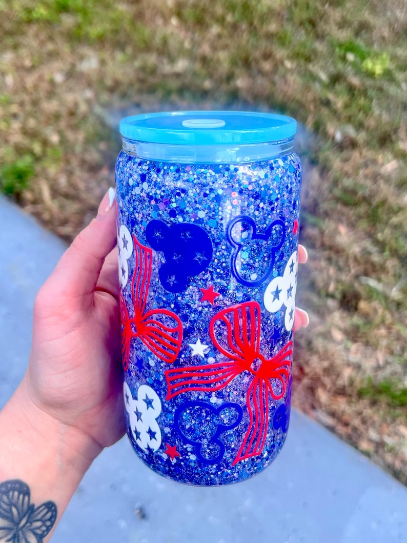 May include: A blue glitter tumbler with a matching blue lid. The tumbler is decorated with red bows, white clouds, blue Mickey Mouse silhouettes, and white stars. The tumbler is held by a person.