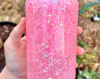 May include: A clear glass tumbler filled with pink glitter and iridescent specks. The tumbler has a bamboo lid. The glass is held by a hand, with a blurred outdoor background.