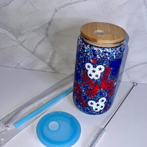 May include: A glass tumbler with a wooden lid, blue lid, straw, and cleaning brush. The tumbler features red bows and white stars on a blue glitter background. Designed for cold drinks, it has a capacity of approximately 470 ml.