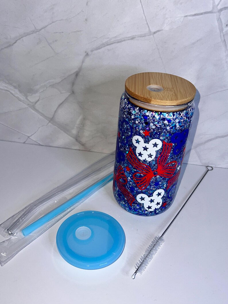 May include: A glass tumbler with a wooden lid, blue lid, straw, and cleaning brush. The tumbler features red bows and white stars on a blue glitter background. Designed for cold drinks, it has a capacity of approximately 470 ml.