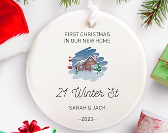 May include: White ceramic Christmas ornament with a winter scene of a cabin, a lighthouse, and snow-covered trees. The text reads "First Christmas in our new home 21 Winter St Sarah & Jack -2023-"