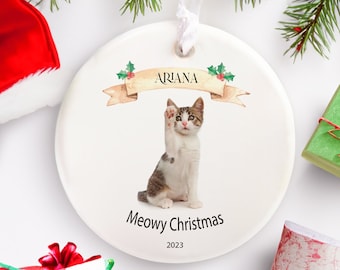 May include: White ceramic Christmas ornament with a cat wearing a red collar and raising its paw. The ornament has a banner with the name "Ariana" and the text "Meowy Christmas 2023".