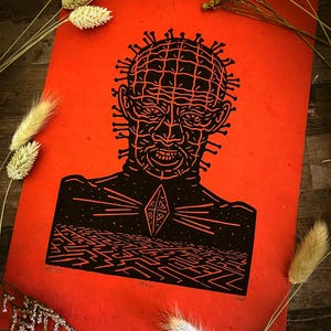 Puede incluir: Una impresión roja con una ilustración negra de una figura con alfileres en la cabeza y un diseño geométrico en el pecho. Debajo, un patrón laberíntico. La palabra "Hell" está impresa en la parte inferior derecha.