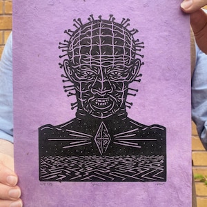 May include: A black and white linocut print on lavender paper. The print features a detailed portrait of a character with pins in their head, a diamond shape on their chest, and a maze at the bottom. The print is signed and numbered.