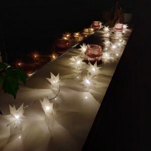 Könnte beinhalten: Eine Lichterkette aus weißen Papiersternen mit warmweißen LED-Lampen. Die Lichter sind über eine weiße Oberfläche gespannt und spiegeln sich in einem Spiegel hinter ihnen.