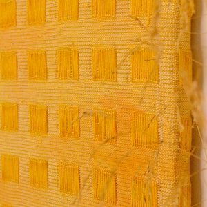 May include: A yellow woven wall hanging with a grid pattern. The hanging is made of a textured fabric with a subtle ombre effect.