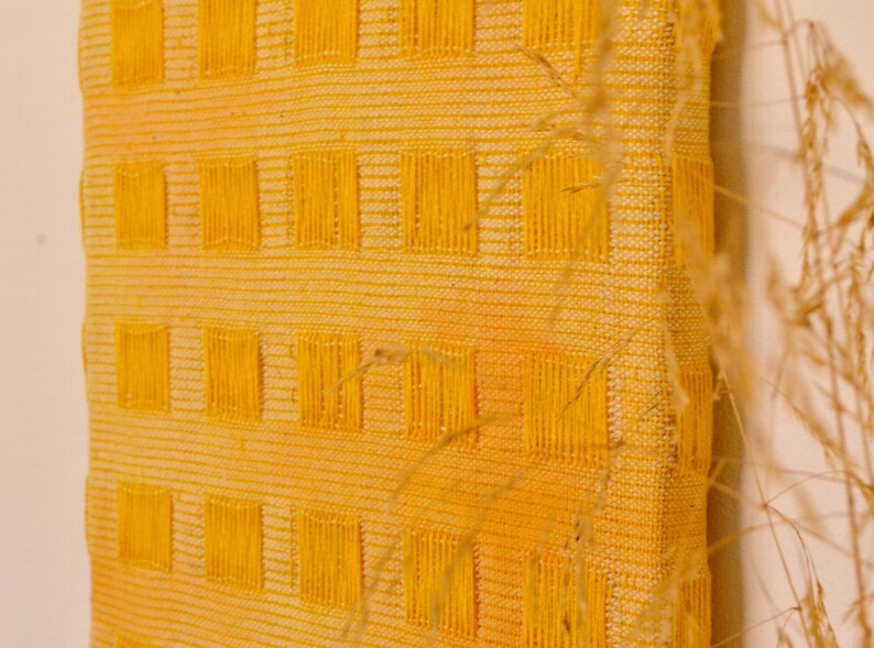 May include: A yellow woven wall hanging with a grid pattern. The hanging is made of a textured fabric with a subtle ombre effect.