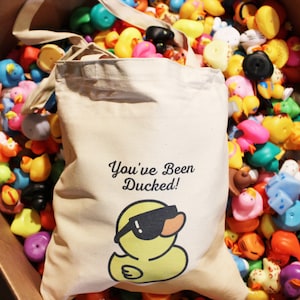May include: A white canvas tote bag with the text "You've Been Ducked!" and a cartoon duck wearing sunglasses. The bag is surrounded by a large pile of colorful rubber ducks.