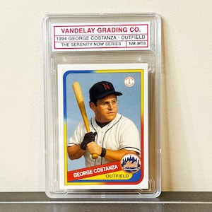 May include: A graded baseball card featuring George Costanza in a Mets uniform, holding a bat. The card is encased in a clear protective holder. Text on the card reads "Vandelay Grading Co." and "The Serenity Now Series."