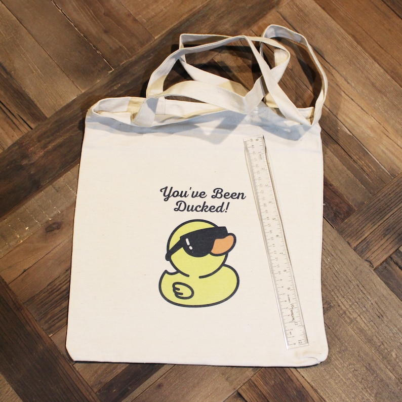 May include: A white canvas tote bag with a yellow rubber duck wearing sunglasses and the text "You've Been Ducked!" printed on it.