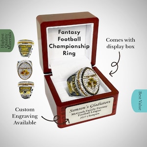 May include: A championship ring for fantasy football, displayed in a wooden box. The ring is silver and gold, encrusted with crystals, and features the text "CHAMPION". The box is engraved with "Samson's Gladiators" and "2025 Champion".