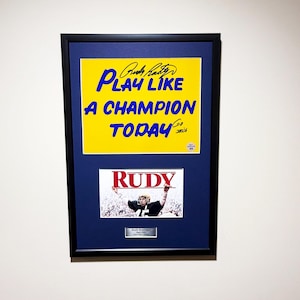 May include: Framed artwork featuring the text "PLAY LIKE A CHAMPION TODAY" in blue on a yellow background, with a signature. Below is a photo of a football player holding a sign that says "RUDY".