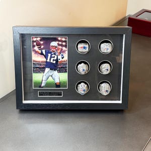 May include: A framed display featuring a photo of a football player and six championship rings. The rings are silver-toned with intricate detailing. The photo shows the player in a blue and white uniform. The frame is black with a white border.