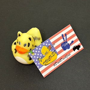 May include: Yellow rubber duck with a leopard print design and a green leaf on its head. The duck is sitting on a red, white, and blue American flag with the text "You've Been DUCKED" and the hashtag "#DuckDuckJeep".