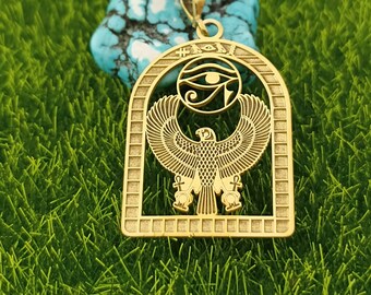 May include: Gold-colored pendant necklace featuring an Egyptian design. The pendant showcases a stylized eagle with outstretched wings, the Eye of Horus, and hieroglyphic symbols. The necklace is displayed on a turquoise stone against a backdrop of green grass.
