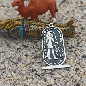 Könnte beinhalten: Eine silberne Halskette mit Anhänger und ägyptischem Hieroglyphen-Design. Der Anhänger hat einen dunklen Hintergrund mit einer detaillierten silbernen Figur und Symbolen. Eine orangefarbene Kamel-Figur und ein dekorierter Sarkophag befinden sich im Hintergrund.