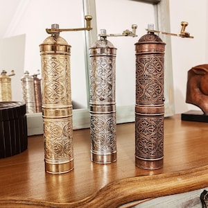 Puede incluir: Tres molinillos de pimienta de estilo antiguo con diseños intrincados. Los molinillos están hechos de metal y tienen un acabado dorado, plateado y cobre.