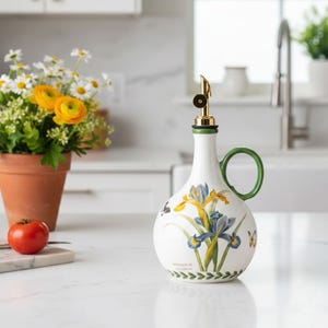 May include: White ceramic oil bottle with a green handle and gold spout. The bottle is decorated with blue and yellow iris flowers and butterflies. A tomato and a pot of flowers are in the background.