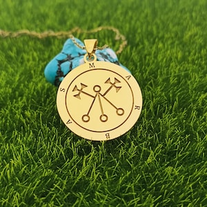 May include: A gold pendant necklace with a circular charm featuring a symbol with four arms and a circle in the center. The charm is inscribed with the letters "MARS" around the edge.