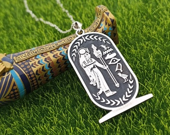 May include: Silver pendant necklace with an Egyptian hieroglyphic design. The pendant features a figure of a pharaoh holding a staff and an ankh symbol. The pendant is surrounded by a decorative border.