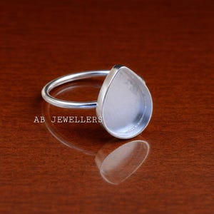 Op de afbeelding: Een zilveren ring met een druppelvormige zetting. De ring ligt op een bruin oppervlak. De tekst "AB JEWELLERS" staat op het oppervlak.