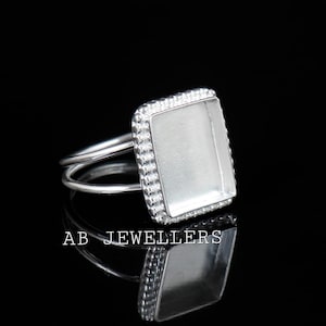 May include: A silver-toned ring featuring a rectangular bezel setting with a beaded edge. The band is split into two thin, parallel wires that connect to the sides of the setting. The text AB JEWELLERS is visible below the ring.