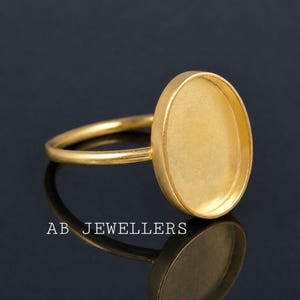 Puede incluir: Un anillo de tono dorado con un engaste ovalado liso diseñado para un cabochon o incrustación. El anillo tiene una banda delgada y redondeada con un acabado mate. El texto AB JEWELLERS está impreso en blanco cerca de la base del anillo.