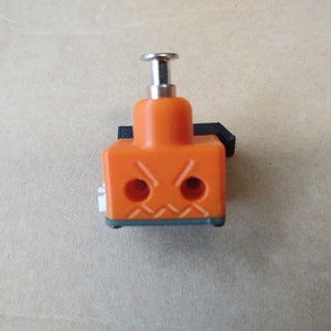 Puede incluir: Válvula solenoide de plástico naranja y negro con un botón pulsador plateado en la parte superior. La válvula tiene dos puertos redondos en la parte delantera.