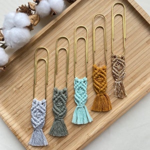 May include: Five gold paper clips with colorful macrame tassels attached. The tassels are in shades of gray, green, blue, yellow, and brown.