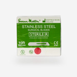 Könnte beinhalten: Eine grün-weiße Packung mit 10 sterilen chirurgischen Klingen aus Edelstahl. Die Packung ist mit "Swann-Morton®" und "STERILE R" beschriftet.