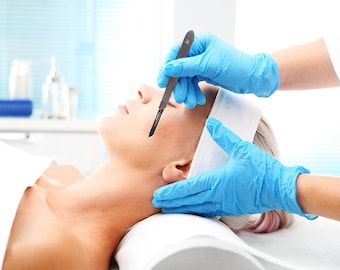 May include: A person lying on a white table with a white towel on their head. A gloved hand holds a scalpel over their face.
