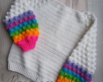 Puede incluir: Suéter blanco de ganchillo con mangas arcoíris. Las mangas presentan un vibrante patrón de rosa, naranja, amarillo, verde, azul y morado. El cuerpo del suéter es de color blanco liso.