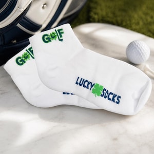 May include: A pair of white ankle socks with the word "GOLF" in green and the words "LUCKY SOCKS" in blue and green. A golf ball and tee are in the background. The socks are on a marble surface.