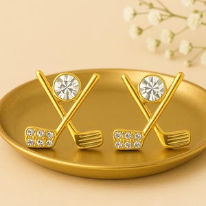 May include: A pair of gold-tone earrings with a golf club and ball design. Each earring features a clear crystal stone in the center of the design.
