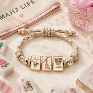 May include: A braided cord bracelet featuring three rectangular charms with enamel illustrations of a joker, a bird on a bottle, and a lion head with the text BAM. The bracelet includes gold-toned metal beads and adjustable knots.