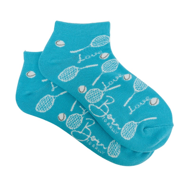Puede incluir: Un par de calcetines tobilleros turquesa con tem&aacute;tica de tenis. Los calcetines presentan un estampado de raquetas de tenis blancas, pelotas de tenis y las palabras "Love" y "Born to Rally" en una fuente de escritura. Los calcetines son de material suave.