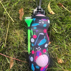 May include: A black golf ball holder with a colorful golf-themed pattern, including golf clubs, balls, and flowers. A green golf tee is attached. A pink golf ball is visible in the holder.