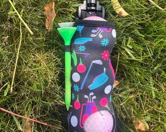May include: A black golf ball holder with a colorful golf-themed pattern, including golf clubs, balls, and flowers. A green golf tee is attached. A pink golf ball is visible in the holder.