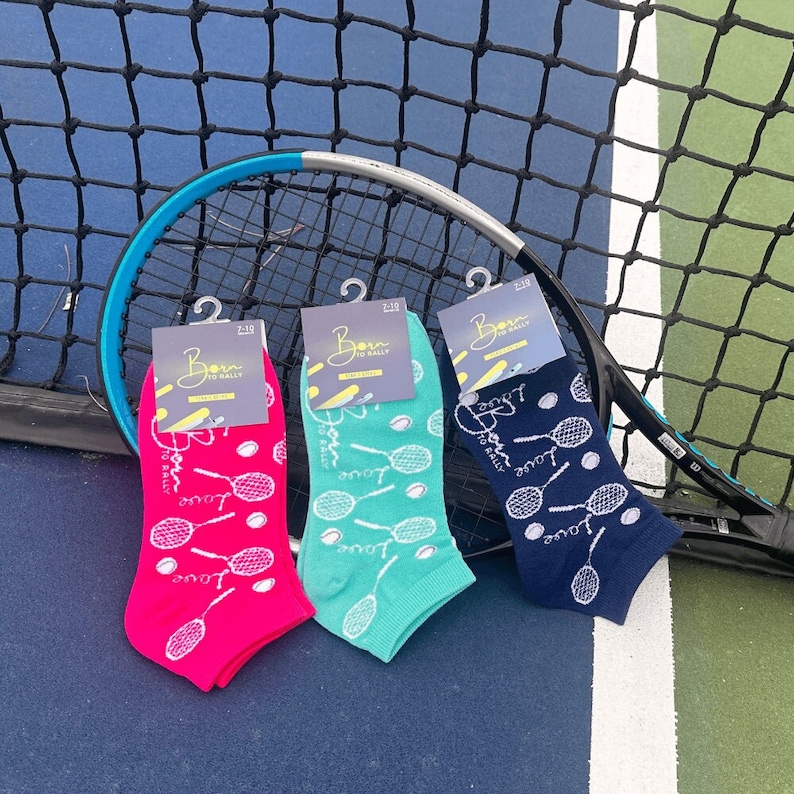 Puede incluir: Tres pares de calcetines tobilleros en rosa, verde azulado y azul marino, cada uno con un estampado de raqueta y pelota de tenis. Los calcetines se exhiben con una raqueta de tenis sobre un fondo de cancha de tenis. Cada calcet&iacute;n tiene una etiqueta que dice "Born to Rally".