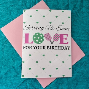 K&ouml;nnte beinhalten: Eine wei&szlig;e Karte mit einem Muster aus gr&uuml;nen und rosa Herzen. Die Karte sagt "Serving Up Some LOVE FOR YOUR BIRTHDAY" mit einem gr&uuml;n-wei&szlig;en Pickleball und zwei rosa Pickleball-Schl&auml;gern.