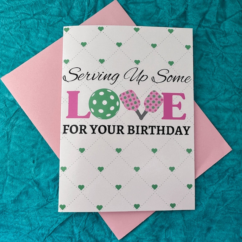 K&ouml;nnte beinhalten: Eine wei&szlig;e Karte mit einem Muster aus gr&uuml;nen und rosa Herzen. Die Karte sagt "Serving Up Some LOVE FOR YOUR BIRTHDAY" mit einem gr&uuml;n-wei&szlig;en Pickleball und zwei rosa Pickleball-Schl&auml;gern.