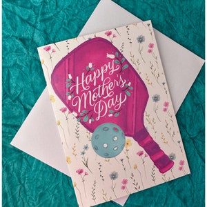 K&ouml;nnte beinhalten: Eine rosa und wei&szlig;e Gru&szlig;karte mit einem Blumenmuster. Die Karte zeigt einen rosa Schl&auml;ger mit dem Text "Happy Mother's Day" in wei&szlig;er Schrift.