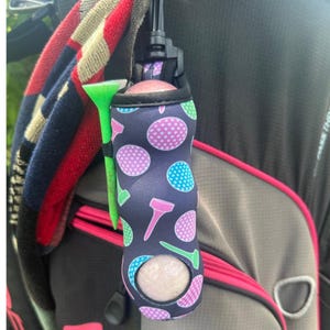 May include: A navy blue golf ball holder with a pattern of pink, purple, and blue golf balls and tees. A green golf tee is attached to the holder. A golf ball is visible inside the holder.