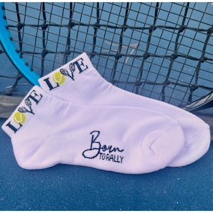 Puede incluir: Un par de calcetines tobilleros de color lavanda con la palabra "LOVE" y un gr&aacute;fico de pelota de tenis en el pu&ntilde;o. La frase "Born to Rally" est&aacute; bordada en negro en el lateral. Una raqueta de tenis en el fondo.