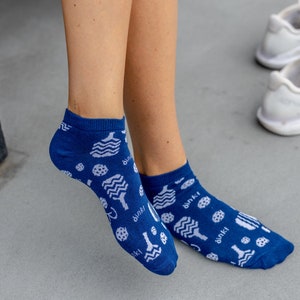 May include: Blue ankle socks with a white pattern of pickleball paddles, balls, and other pickleball-related graphics. The word "dink!" is printed on the socks. The socks are worn by a person.