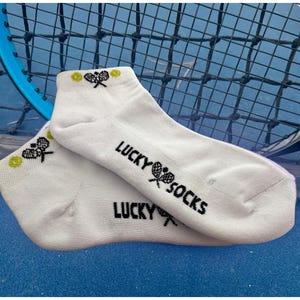 Puede incluir: Calcetines tobilleros blancos con el texto "LUCKY SOCKS" en negro. Los calcetines presentan un dise&ntilde;o de raqueta y pelota de tenis. Una raqueta y una red de tenis son visibles en el fondo.